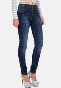 Jeans skinny in denim blu scuro con vita alta, dotati di tasche frontali, cuciture sottili e una texture liscia. Abbinati a scarpe con tacco argentate.