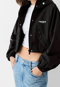 Svart kort jacka med elastiska ärmar, dragkedja fram och justerbar huva. Har en logotyp och två sidofickor. Bärs över en vit topp.