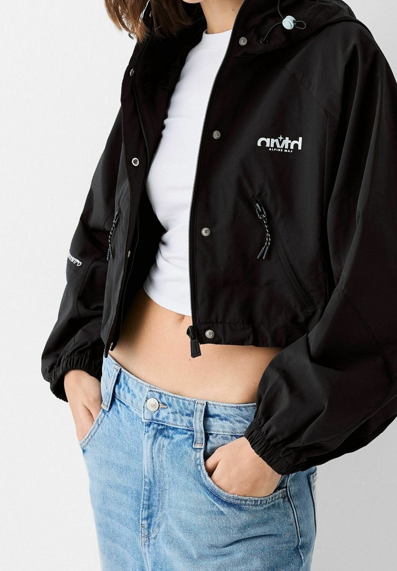 Svart kort jacka med elastiska ärmar, dragkedja fram och justerbar huva. Har en logotyp och två sidofickor. Bärs över en vit topp.