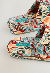 Par af farverige blomstermønstrede bløde hjemmesko med turkis "NEXT" mærker på en lys baggrund.