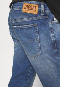 Blue denim jeans with a faded wash. Features a leather "DIESEL" label at the waist and back pockets with contrasting stitching details.