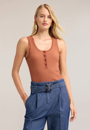 Vrouw met lang blond haar, gekleed in een mouwloos geribd roestkleurig topje en hooggetailleerde blauwe broek met gevlochten riem en gouden armbanden.