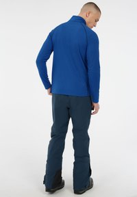 Modrý pulóver s dlhým rukávom a golierom, spárovaný s tmavozelenými nepremokavými nohavicami so bočnými zipsami a čiernymi akcentmi, zobrazený zozadu.