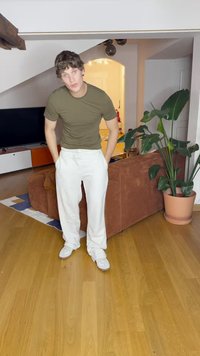 Olive green fitted t-shirt, white pants with pockets, and white sneakers. Background includes a brown couch, potted plant, and TV.