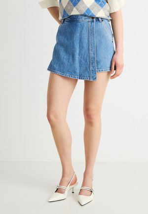 Donna che indossa una minigonna di jeans blu, un maglione a rombi e décolleté slingback bianchi con punte appuntite e cinturini con fibbia.