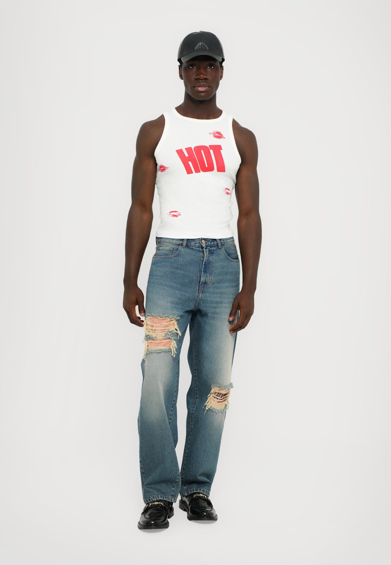 Witte tanktop met "HOT" in rode letters en rode lippenafdrukken, gecombineerd met lichtblauwe ripped jeans. Model draagt een zwarte pet en schoenen.