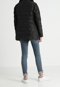 Svart quiltad pufferjacka med hög krage, med horisontell sömnad och avslappnad passform, tillsammans med blå tighta jeans och vita sneakers.