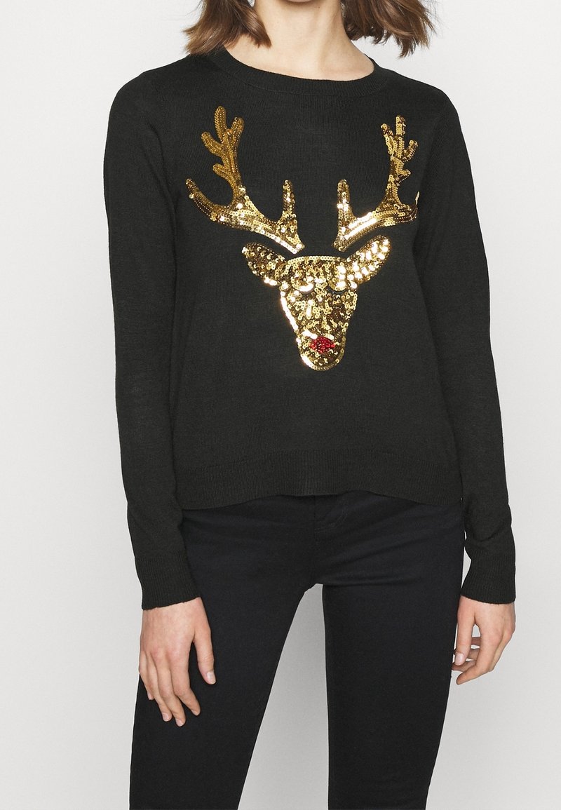 Frau, die einen schwarzen Pullover trägt, der ein mit Pailletten verzierte Rentierkopf mit goldenen Geweih und einer roten Nase zeigt, kombiniert mit dunklen Jeans.