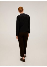 Giacca nera aderente con maniche lunghe, abbinata a pantaloni marroni scuri. Il modello indossa scarpe scure, che mostrano un design semplice ed elegante.