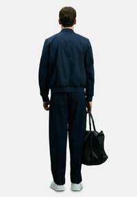 Marineblaue Bomberjacke mit gerippten Bündchen und Saum, kombiniert mit passenden Hosen. Weiße Sneaker und eine schwarze Tote-Bag ergänzen das Outfit.