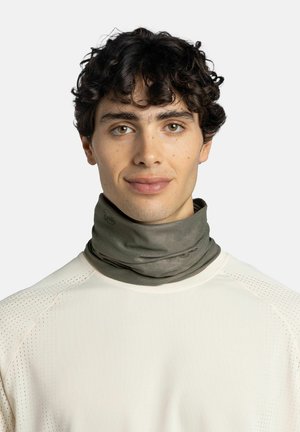 Junger Mann mit lockigem dunklem Haar, der ein beiges perforiertes Langarmhemd und einen olivgrünen Halswärmer vor einem einfarbigen Hintergrund trägt.