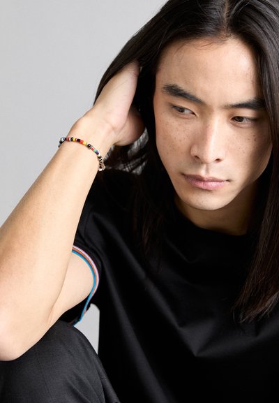 T-shirt noir avec un col rayé multicolore, associé à un bracelet en perles de différentes couleurs. Cheveux longs et foncés visibles.