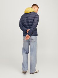 Jūrinis pūkinis švarkas su geltonu kapišonu, pasižymintis siuvinėta tekstūra ir užtrauktuku. Nešiojamas su šviesiai mėlynais džinsais ir tamsiomis šlepetėmis.