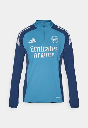 Blå langermet treningstrøye med glidelås i nakken, med Arsenal-logo og teksten "Emirates FLY BETTER", samt marineblå sidestykker og hvite striper.