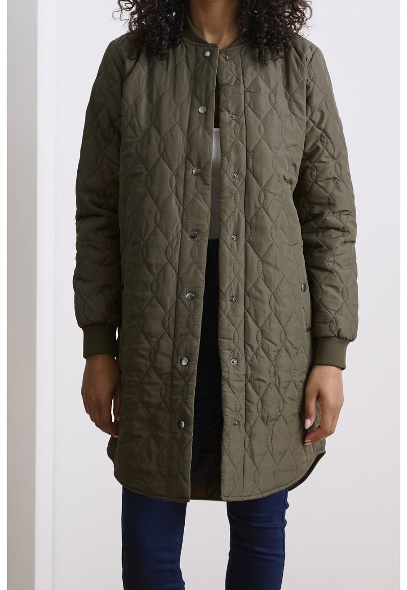 Personne portant une longue veste matelassée vert olive avec des boutons-pression et des poignets côtelés, associée à un pantalon bleu foncé.