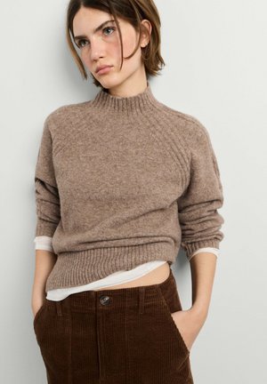Junge Frau mit kurzen Haaren, die einen braunen Strickpullover über einem weißen Hemd und dunkelbraune Cordhosen trägt, mit den Händen in den Taschen.