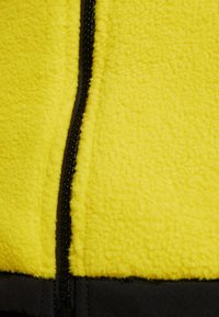 Žlutá fleecová látka s černým zipem a černým lemováním na spodní straně, zobrazující texturu a detaily švů.