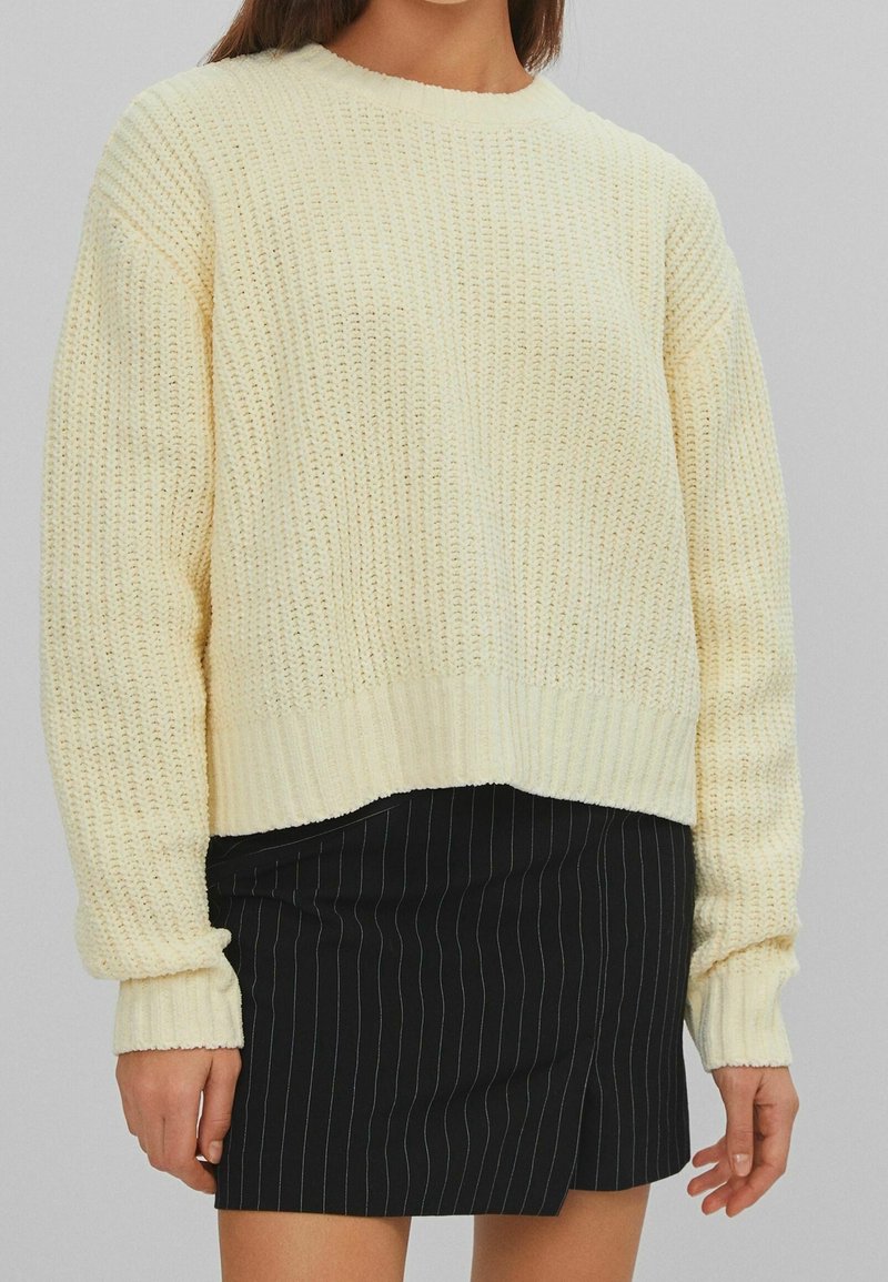 Kremowy sweter z dzianiny o luźnym kroju, z prążkowanymi mankietami i dołem oraz teksturowanym wzorem, zestawiony z czarną, wąsko prążkowaną spódnicą.