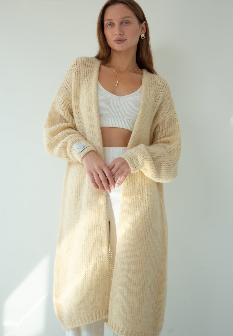 Jasno beżowy kardigan o prążkowanej fakturze, z otwartym przodem i długimi rękawami. Nosi się go na biały top crop i dopasowane spodnie. Miękki, przytulny materiał.
