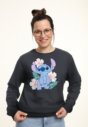 Kvinde med briller og håret sat op i en knold iført en sort sweatshirt med den blå tegneseriefigur Stitch og lyserøde og hvide blomster.