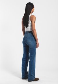 Jeans de tiro alto en azul con pernera acampanada, fabricados en denim. Presentan bolsillos traseros y una etiqueta sencilla en la cinturilla. Combinados con una camiseta sin mangas ajustada blanca.