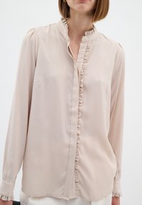Blusa beige chiaro con maniche lunghe, colletto arricciato e chiusura con bottoni. Tessuto liscio con una vestibilità comoda e una lieve lucentezza.