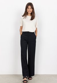 Pantalons noirs ajustés à taille haute, associés à un haut crème à manches courtes ; le modèle porte des sandales noires tressées et se tient sur un sol en béton.