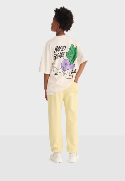 Person iført en cremefarvet oversized T-shirt med teksten "Rapid Radish" og en tegneserie-aftegning af en radise, kombineret med lysegule bukser og hvide sneakers.