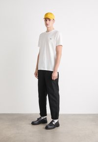 Camiseta de algodón blanca con un pequeño logo, pantalones negros, gorra amarilla, zapatos negros con detalles plateados y calcetines blancos. Atuendo sencillo y casual.
