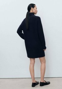 Donna con lunghi capelli scuri raccolti in una coda di cavallo bassa, che indossa un vestito a maglia blu navy e mocassini neri, in piedi contro un muro bianco.