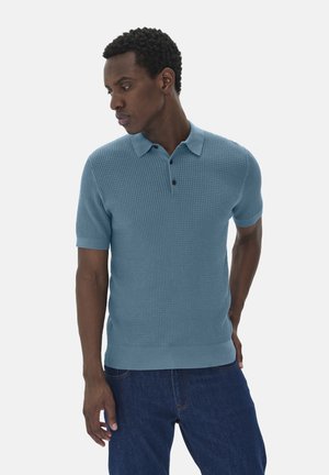 Polo en maille bleu clair à manches courtes, design texturé, col plat et patte de boutonnage à trois boutons, associé à un jean sombre.