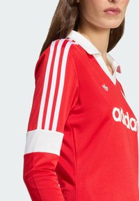 Rød idrettsjakke med hvit krage og mansjetter, med tre hvite striper på ermene og et adidas-logo på brystet.
