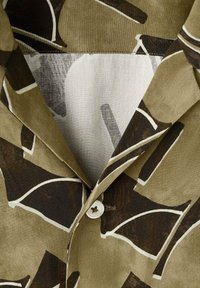 Chemise à manches courtes en vert olive avec des motifs abstraits marron foncé et blancs. Dotée d'un col pointu et d'un bouton blanc à l'avant.