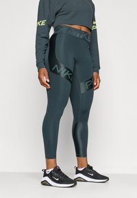 Nike tajice visokog struka u tamnozelenoj boji s glatkom teksturom, sa kontrastnim zelenim logotipom i prilagođenim dizajnom. Crne sportske cipele upotpunjuju izgled.