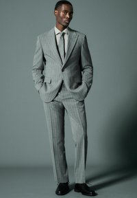 Traje gris de rayas con chaqueta de solapa muesca y pantalones a juego. Acompañado por una corbata gris claro y zapatos de vestir negros.