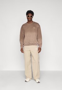Pruun kapuutsiga sweatshirt, sobivad beežid vabalt lõigatud püksid ja valged tossud, mida täiendab heledat värvi päikeseprillid.