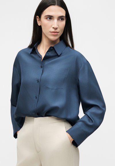 Γυναίκα με μακριά σκούρα μαλλιά που φοράει ένα σκούρο μπλε πουκάμισο με κουμπιά μπαλωμένο σε μπεζ παντελόνια με υψηλή μέση, τα χέρια στις τσέπες, κοιτάζει προς τα πλάγια.