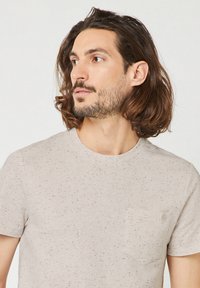 Uomo con capelli castano medio e barba, che indossa una t-shirt beige chiaro a pois con taschino sul petto, che guarda a sinistra.