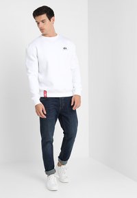 Hvid sweatshirt med rund halsudskæring, ribbede manchetter og kant. Bemærkelsesværdigt sort logo på brystet. Kombination med mørke denimjeans og hvide sneakers.