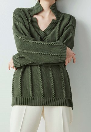 Persona con un suéter de punto verde oliva con patrones verticales texturizados y cuello en V, combinado con pantalones de color crema.