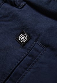 Tessuto blu navy con una finitura texture, caratterizzato da un piccolo logo nero applicato e cuciture visibili nell'area della tasca.