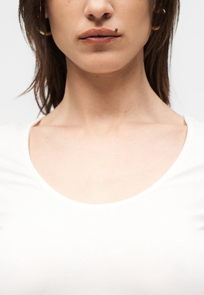 Parte inferior del rostro y cuello de mujer, con camiseta blanca, con un lunar oscuro y prominente sobre el labio superior en el lado derecho.