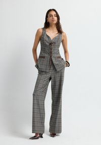 Grijze geruite op maat gemaakte jumpsuit met een aansluitend lijfje, v-halslijn en bruine leren accenten. Heeft wijde benen en een vetersluiting aan de voorkant.