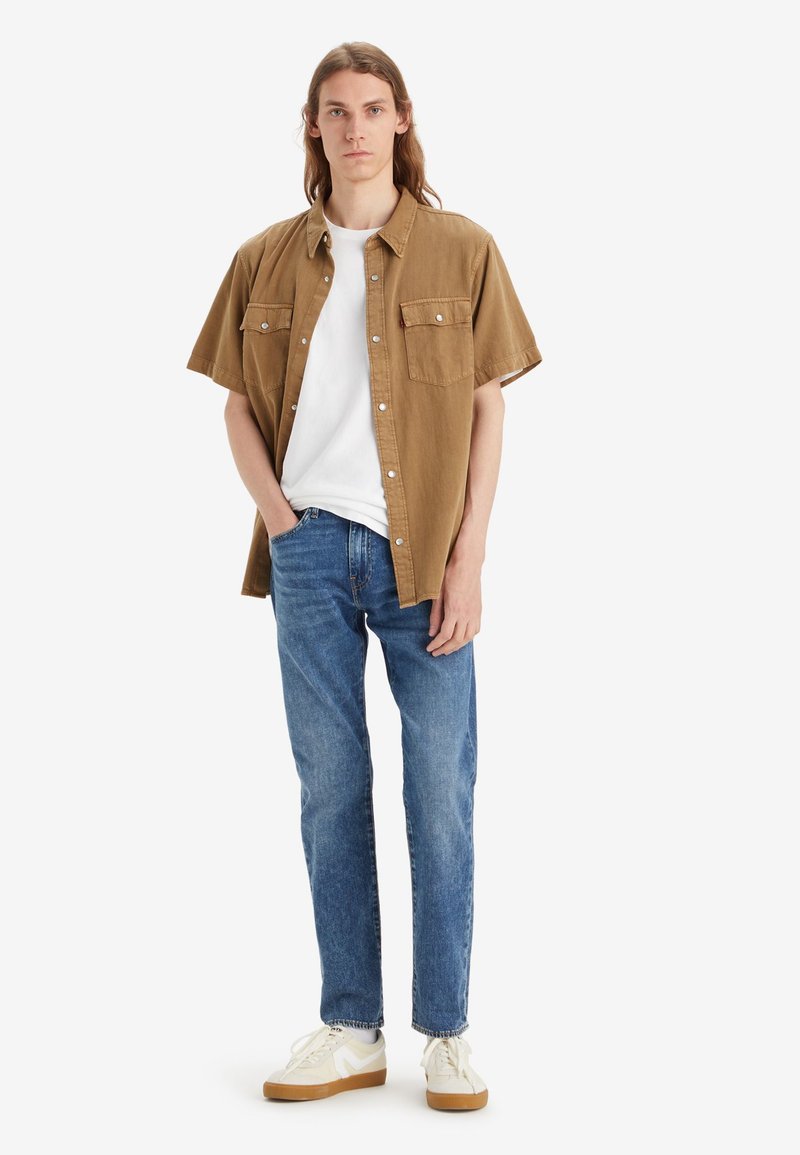 Braunes kurzärmliges Hemd mit Brusttaschen, kombiniert mit einem weißen T-Shirt und blauen Jeans. Die Schuhe sind weiß mit hellbraunen Sohlen.