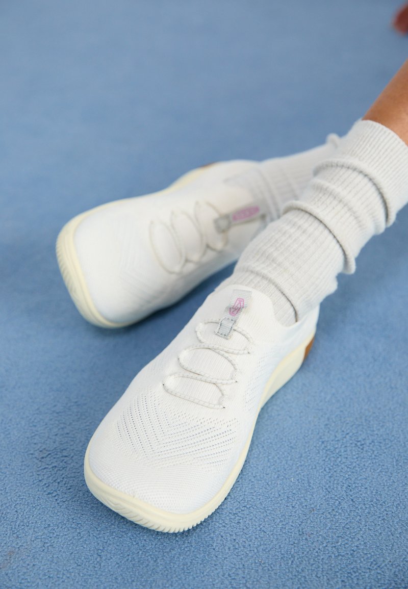 Weiße Sportschuhe mit gemustertem Mesh-Obermaterial, elastischen Schnürsenkeln und Gummisohlen. Paar getragen mit hellgrauen gerippten Socken auf blauer Oberfläche.