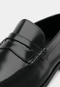 Černý kožený mokasín s hladkým povrchem, ozdobným prošíváním a zaoblenou špičkou. Nárt zdobí dvojité páskové detaily.