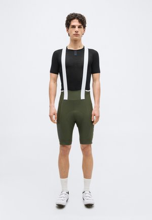 Young man stands wearing black short-sleeve cycling jersey, olive green bib shorts with white straps, white socks, and white cycling shoes.