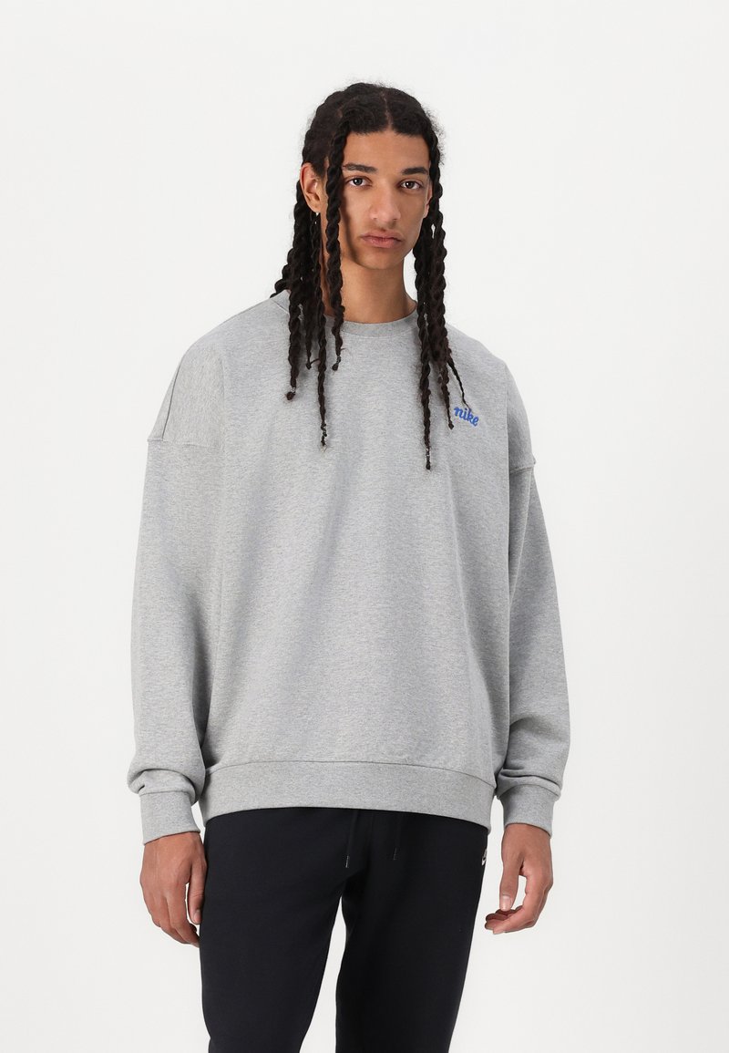 Sweatshirt cinza de gola redonda com um ajuste descontraído, punhos e bainha canelados, apresentando um pequeno logótipo azul no peito. Usado com calças pretas.