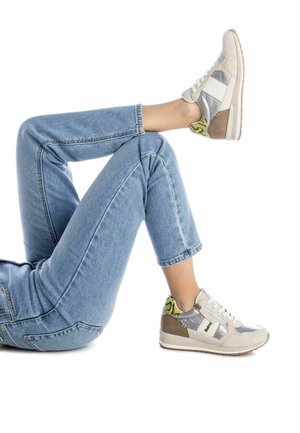 Personne portant un jean bleu clair et des baskets argentées et beiges avec un détail de motif serpent jaune, une jambe pliée, sur fond blanc.