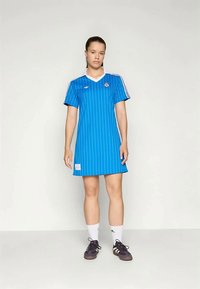 Modra športna obleka s črtami in kratkimi rokavi, bela V-izreza in detajl s logotipom. Kombinirana z belimi nogavicami in temnimi čevlji.